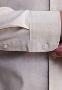 Chemise à manches longues beige clair avec tissu texturé, col à boutons et poignets boutonnés avec bordure cousue.