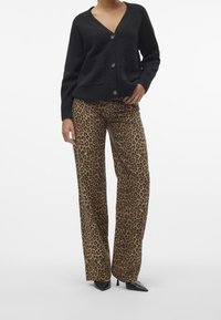 Cardigan en maille noire avec col en V et deux poches avant, associé à un pantalon large à imprimé léopard marron et noir, porté avec des talons noirs.