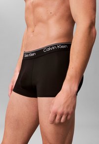 Nærbillede af en mands torso iført sorte Calvin Klein boxershorts med logo i taljebåndet, mod en ensfarvet grå baggrund.