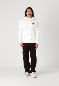 Weißer Hoodie mit einer Fronttasche, Kapuze mit Kordelzug und navy Logo. Kombiniert mit dunkelbraunen Hosen und weißen Sneakers, stehend vor einem einfarbigen Hintergrund.