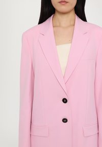 Roze blejzer od glatkog materijala, s rever ovratnikom, dva prednja džepa i dva crna dugmeta. Jednostavan i stručno oblikovan dizajn.