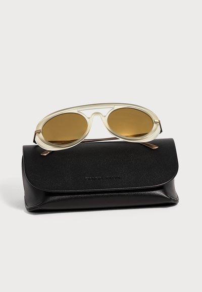 Lunettes de soleil transparentes à monture beige avec verres marron posées sur un étui en cuir noir Giorgio Armani sur un fond clair.
