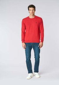 Maglione rosso a maniche lunghe con scollatura rotonda, realizzato in materiale morbido, abbinato a jeans blu e sneakers bianche. Accento del logo nero sul maglione.