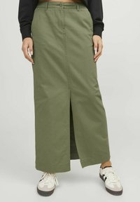 Falda larga de mezclilla verde oliva con abertura frontal, cierre de botón y cremallera, y presillas para cinturón. Combinada con zapatillas blancas y negras.