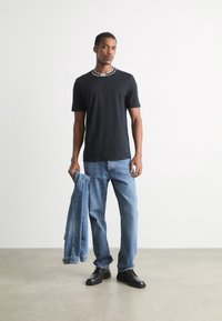 Vyras, vilkintis juodą marškinėlį, mėlynas džinsus ir juodus batus, vienoje rankoje laiko mėlyną džinsinį švarką, stovintis prieš paprastą baltą sieną.