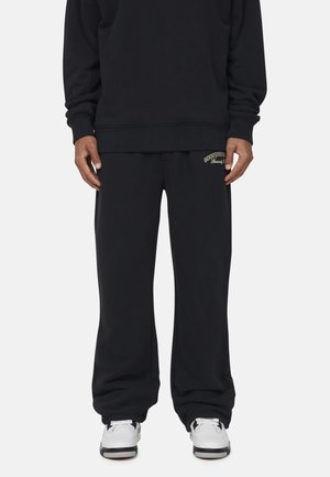Felpa nera con pantaloni sportivi larghi abbinati, con la scritta "GOODSPORT" ricamata in oro sul lato sinistro e sneaker bianche.