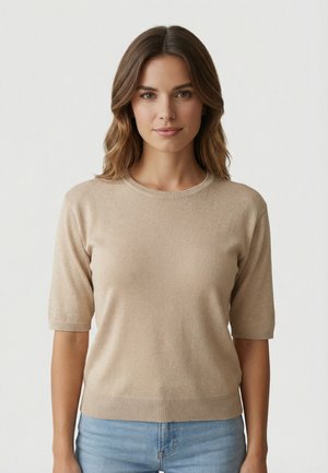 Femme aux cheveux bruns mi-longs portant un haut en maille beige à manches courtes et un jean bleu clair, debout devant un fond clair uni.