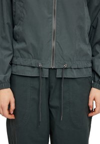 Lightweight, dark green jacket with a zip front, elastic cuffs, and adjustable drawstrings at the hem. Smooth texture and simple design.