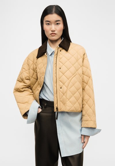 Γυναίκα που φοράει μπεζ καπιτονέ cropped μπουφάν πάνω από μπλε ριγέ πουκάμισο και μαύρο δερμάτινο παντελόνι, στέκεται με το ένα χέρι στην τσέπη.