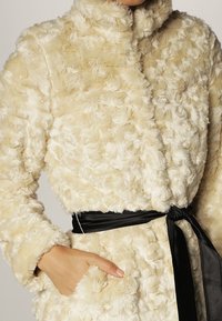 Cremefarvet fake fur-frakke med en tekstureret, fluffy overflade, høj krave og et sort satinbælte i taljen.