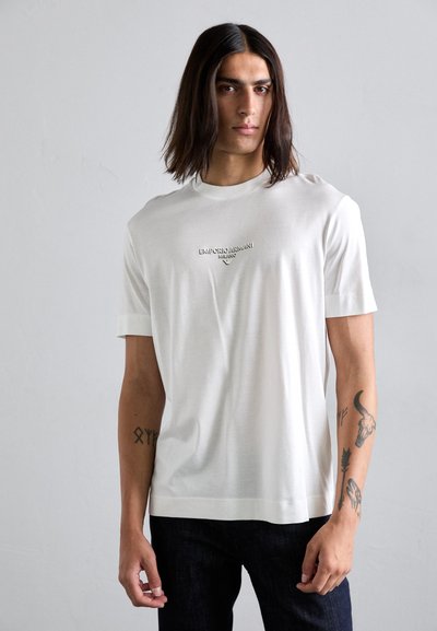 Young man with long dark hair wearing a white Emporio Armani t-shirt and dark jeans, showing tattoos on both forearms.
