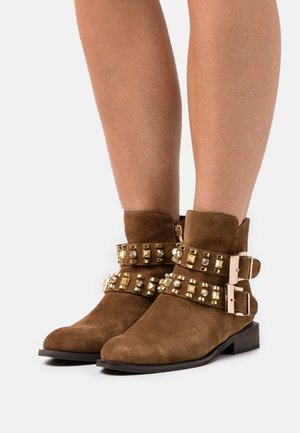 Botines de ante marrón con acentos dorados de tachuelas y dobles hebillas. El diseño presenta un tacón bajo y una puntera redondeada.