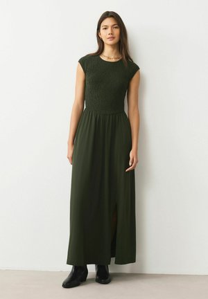 Dunkelgrünes Maxikleid mit gerafftem Oberteil, kurzen Ärmeln und einem Frontschlitz. Kombiniert mit schwarzen Stiefeletten für einen Kontrast. Glatte Textur.