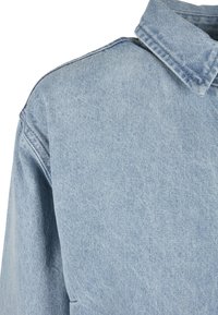 Veste en jean bleu clair en gros plan montrant l'épaule, le col et le haut de la manche avec des coutures visibles et la texture du tissu.
