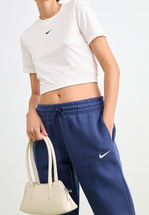 Osoba nosząca białą krótką koszulkę Nike i niebieskie spodnie dresowe Nike, trzymająca małą beżową torebkę na rękę z dwoma uchwytami.