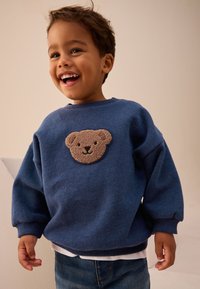 Kék pulóver puha anyagból, az elején barna, texturált medvefolttal. Bordázott mandzsettákkal és laza szabással.