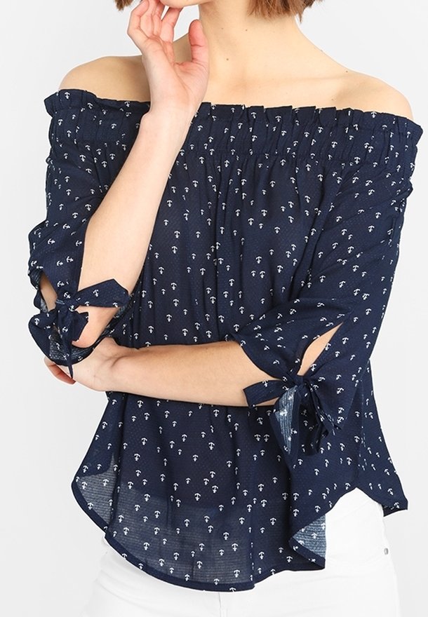 Donna che indossa una blusa navy off-the-shoulder con una piccola stampa di ancore bianche e maniche a 3/4 con fiocchi, abbinata a pantaloni bianchi.