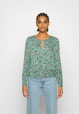 Blusa floral verde com mangas longas, decote em lágrima, cintura franzida e padrão floral multicolorido em tecido suave. Usada com calças de ganga.