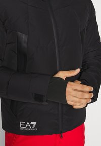 Svart isolerad jacka med dragkedjestängning, texturerat tyg, kardborrejusterbara ärmslut och EA7-logotyp. Modellen bär röda byxor.