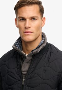 Veste matelassée noire avec col montant, dotée d'une doublure intérieure à carreaux. Fermeture éclair et détails en tissu texturé. Logo subtil.