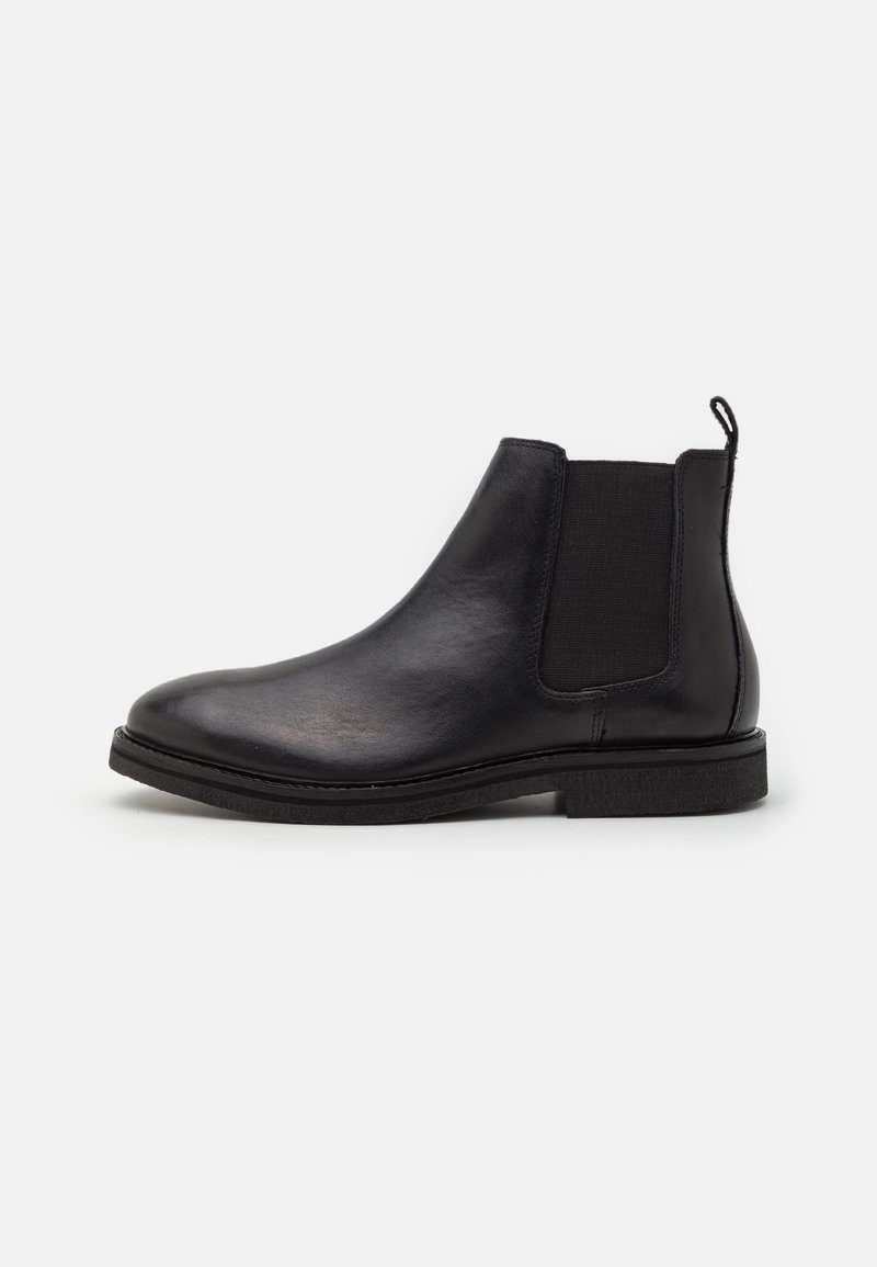 Bottine Chelsea en cuir noir avec panneau latéral élastique, languette à l'arrière et petit talon empilé sur fond blanc.