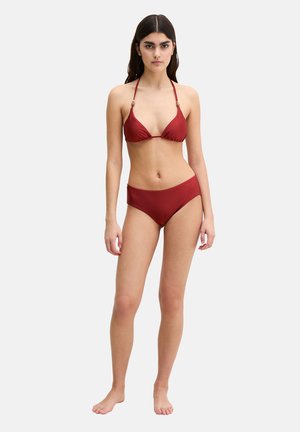 Junge Frau steht barfuß und trägt einen roten Bikini mit Neckholder-Oberteil und mittelhohem Slip vor einem schlichten, hellen Hintergrund.