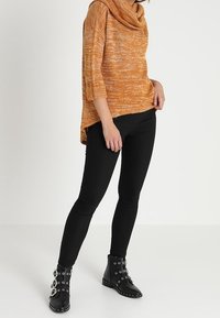 Woman wearing orange textured cowl-neck sweater, black slim pants, and black ankle boots with buckles and studs, standing against white background.