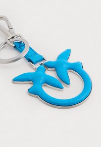 Chaveiro de couro azul com duas formas de pássaros, uma base circular e um acabamento em metal prateado brilhante. Textura suave com cor vibrante.