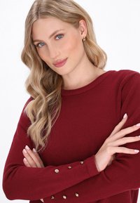 Burgunderfarbener Pullover mit langen Ärmeln, rundem Ausschnitt und goldenen Knopfdetails an den Bündchen. Weiche Textur und taillierte Passform.
