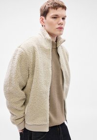 Bež flis jakna s teksturiranom površinom, visokim ovratnikom i zatvaračem. Nošena preko svijetlosmeđeg pletenog džempera i tamnih traperica.