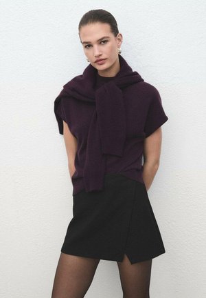 Maglione a maniche corte di un profondo viola con un design di sciarpa annodata sopra una mini gonna nera, abbinato a collant neri trasparenti su uno sfondo bianco.