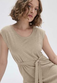 Beige Kleid mit kurzen Ärmeln aus weichem Stoff und lässiger Passform, mit einem Taillenband für anpassbare Stylingmöglichkeiten und subtiler Textur.