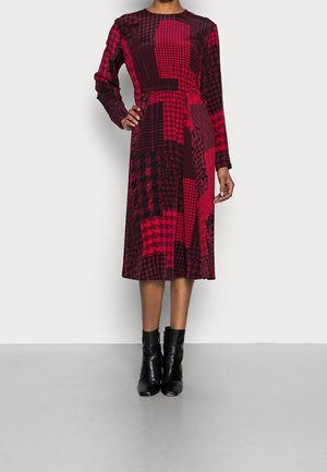 Long-sleeve midi dress with red and black patchwork houndstooth pattern, worn with black ankle boots, standing on a gray floor.