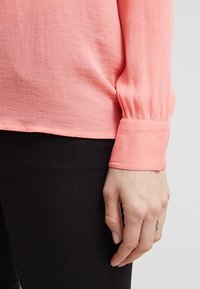 Blusa corallo a maniche lunghe con polsini arricciati, tessuto leggero, orlo dritto e una sottile texture. Abbinata a pantaloni neri attillati.