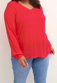Blouse rouge à manches longues avec col en V, en tissu lisse. Le modèle présente une texture subtile et une coupe ample, associé à un jean bleu clair.
