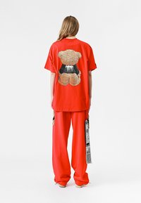 Camiseta roja de manga corta con un gráfico de oso de peluche en la parte de atrás, combinada con pantalones rojos de corte holgado. El material parece suave y ligero.