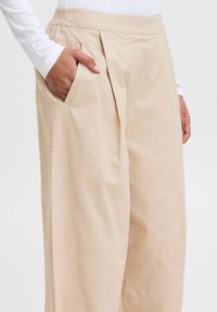 Pantaloni beige a gamba larga realizzati in tessuto leggero, con frontale plissettato e tasche laterali. Texture liscia con vita elastica.