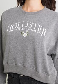 Grå sweatshirt med ribbade cuffar och fåll, som har vit text och blommig grafik. Avslappnad passform med en mjuk, strukturerad yta.