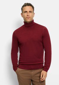 Burgunderfarbener Rollkragenpullover in weichem, geripptem Material, mit langen Ärmeln und figurbetonter Passform, kombiniert mit hellbraunen Hosen.