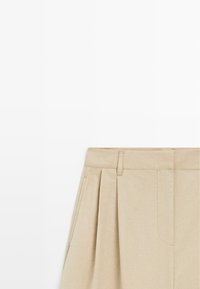 Beżowa plisowana spódnica z szlufkami na pasek i bocznymi kieszeniami, wykonana z gładkiego materiału, pokazana w przyciętym kadrze na białym tle.