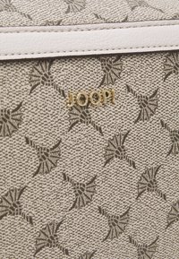 Beige handtas met een verhoogd bloemenpatroon. Voorzien van een gouden "JOOP!" logo en een witte textuurleeraccent.
