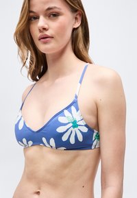 Νεαρή γυναίκα που φοράει μπλε τοπ μπικίνι με λευκά σχέδια μαργαρίτας, κοιτώντας ελαφρώς προς το πλάι, πάνω σε ένα απλό φωτεινό φόντο.
