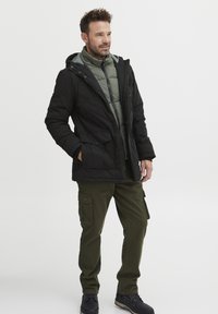 Črna, topla jakna z zelenim notranjim podloženjem, cargo hlače in temni čevlji. Ima kapuco in žepe za funkcionalnost.