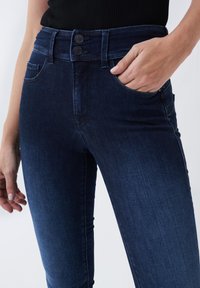 Jeans de mezclilla de color azul oscuro con una cintura alta, cierre de dos botones y bolsillos frontales. La tela presenta una textura suave con un ligero desvanecimiento.
