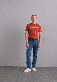 Bărbat stând în picioare împotriva unui perete simplu, purtând un tricou rust-aranj cu sigla Emporio Armani, blugi albaștri, curea neagră și adidași albi.