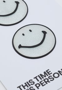 Dois autocolantes redondos de face sorridente metálicos com uma superfície brilhante, contornados a preto, sobre um fundo branco com texto bold a preto em baixo.