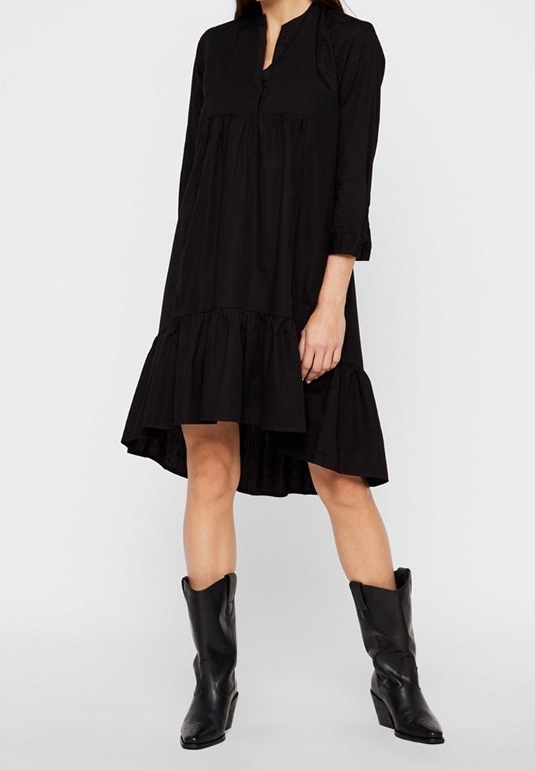 Femme portant une robe noire ample longueur genou avec des manches trois-quarts et des bottes en cuir noires mi-mollet, debout devant un fond uni.