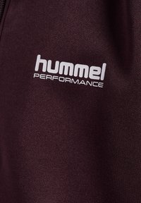 Bordowy materiał o gładkiej teksturze, z logo "hummel PERFORMANCE" w białym kolorze. Zbliżenie na detal odzieży sportowej.