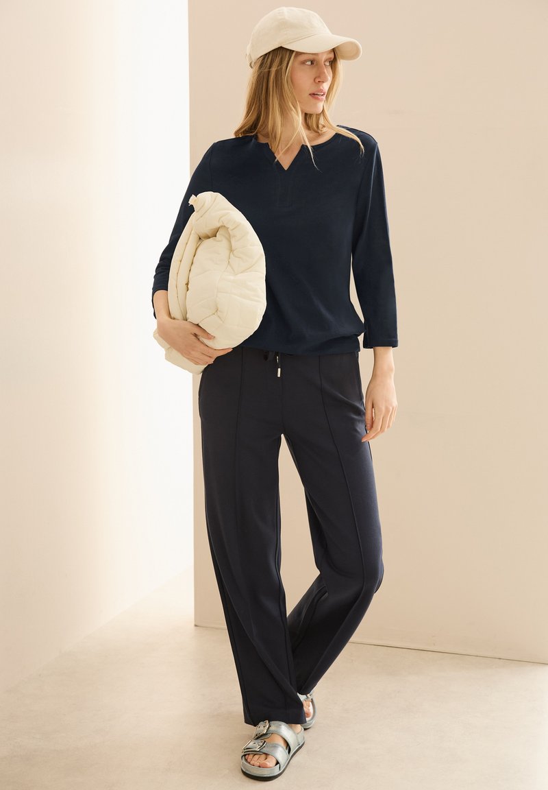 Navyblaues Langarmshirt mit V-Ausschnitt, kombiniert mit dunklen Hosen. Das Model hält eine cremefarbene gesteppten Jacke und trägt metallische Sandalen.