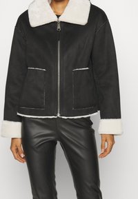 Modell som bär en svart jacka med vit pälskrage, i kombination med svarta läderbyxor, står mot en enkel bakgrund.